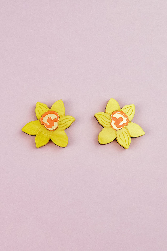 Two yellow Daffodil Stud Earrings with orange centers, resembling, are arranged side by side on a light pink background—perfect for stylish women seeking unique accessories.