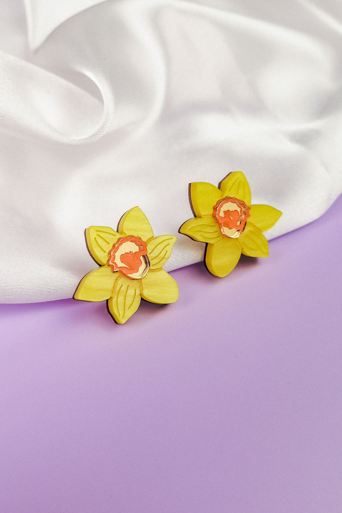 Two yellow Daffodil Stud Earrings with orange centers rest on soft white fabric, set against a pastel purple background—unique accessories perfect for stylish women seeking a floral touch.