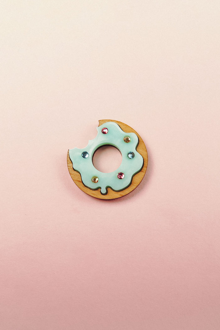 A handcrafted Donut Pin featuring a wooden base, light blue icing, gem-like rhinestones, and a bite taken out, set against a pastel pink background.