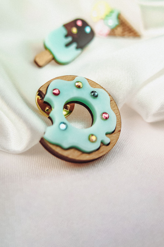 A handcrafted wooden Donut Pin with light blue icing and sparkling rhinestones rests on white fabric. In the background, blurred pins shaped like an ice cream cone and a popsicle add a playful touch.