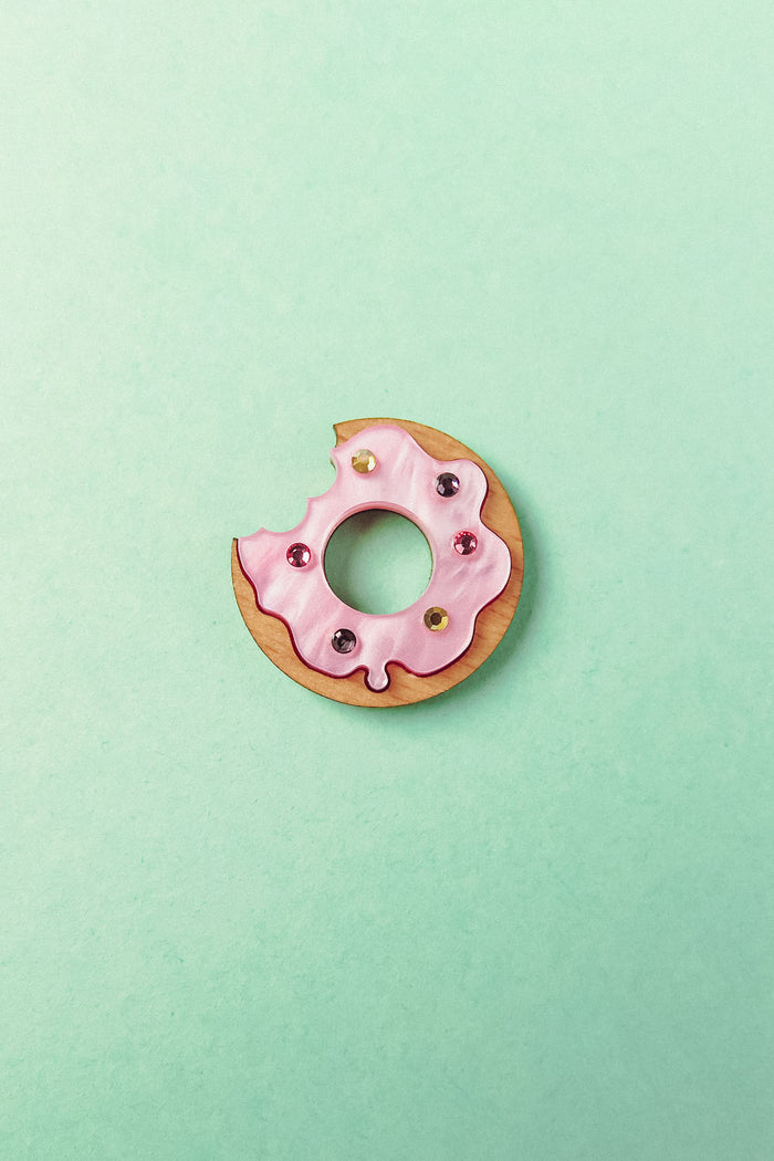 A handcrafted Donut Pin shaped like a donut with pink icing and colorful sprinkles, featuring a bite taken out of it, sits on a pastel mint green background.