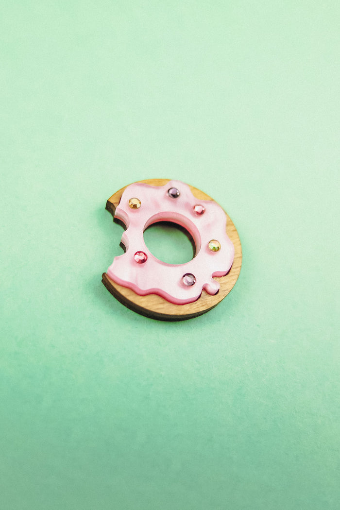 A handcrafted wooden Donut Pin with pink icing and colorful rhinestones, featuring a bite mark, sits on a mint green background.