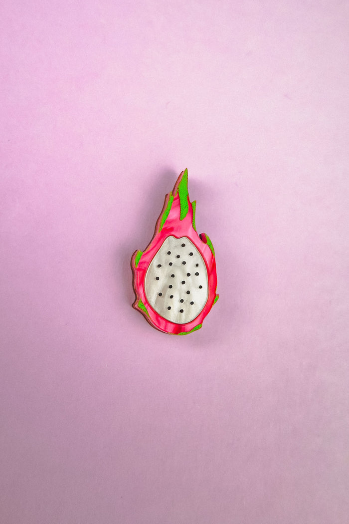 A handcrafted acrylic lapel pin that looks like a halved dragon fruit, with white center, black seeds, and pink and green edges, brings tropical charm against a light purple background.