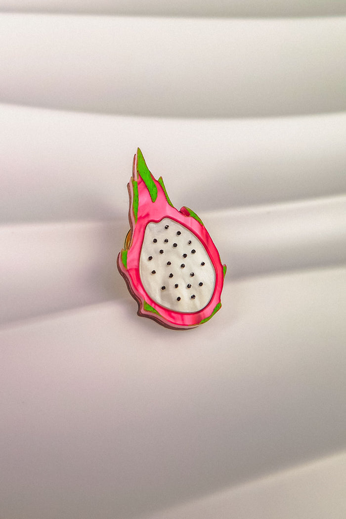 A colorful Dragon Fruit Pin, handcrafted with a white center and black seeds, is surrounded by a pink and green rind, bringing tropical charm to any outfit. Displayed against a wavy, white background.