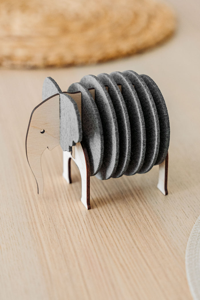 A set of round, gray Elephant Coasters is stacked vertically in a wooden holder shaped like an elephant, adding witty design and furniture protection to your light wood table.