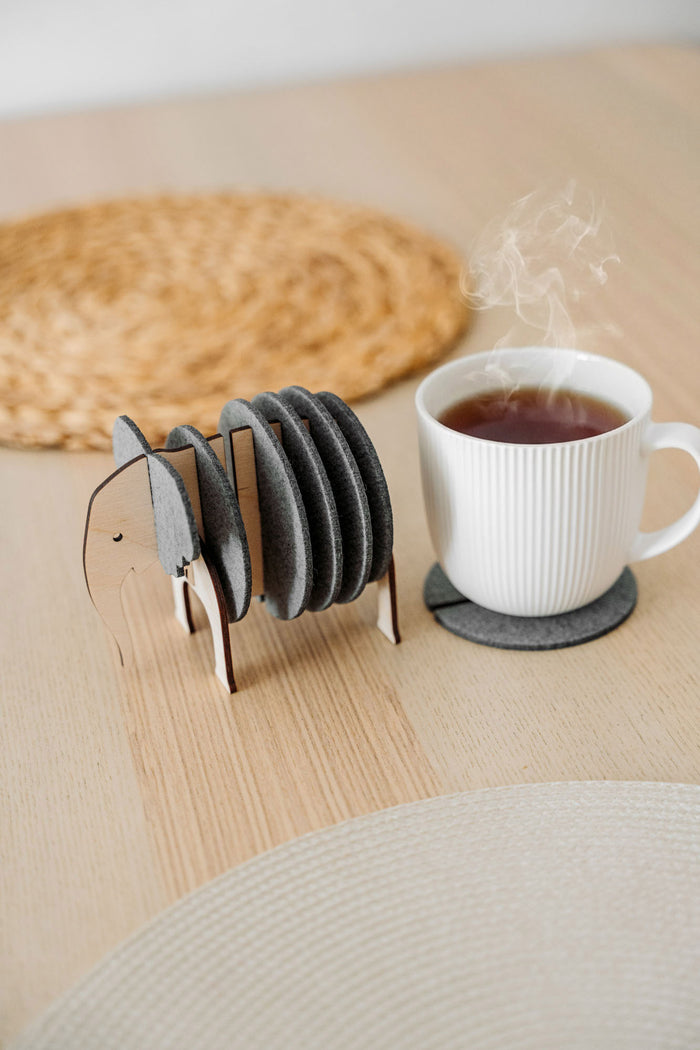 A set of round Elephant Coasters with witty design sits in a wooden elephant-shaped holder on a table, offering furniture protection while a white mug rests on one coaster.