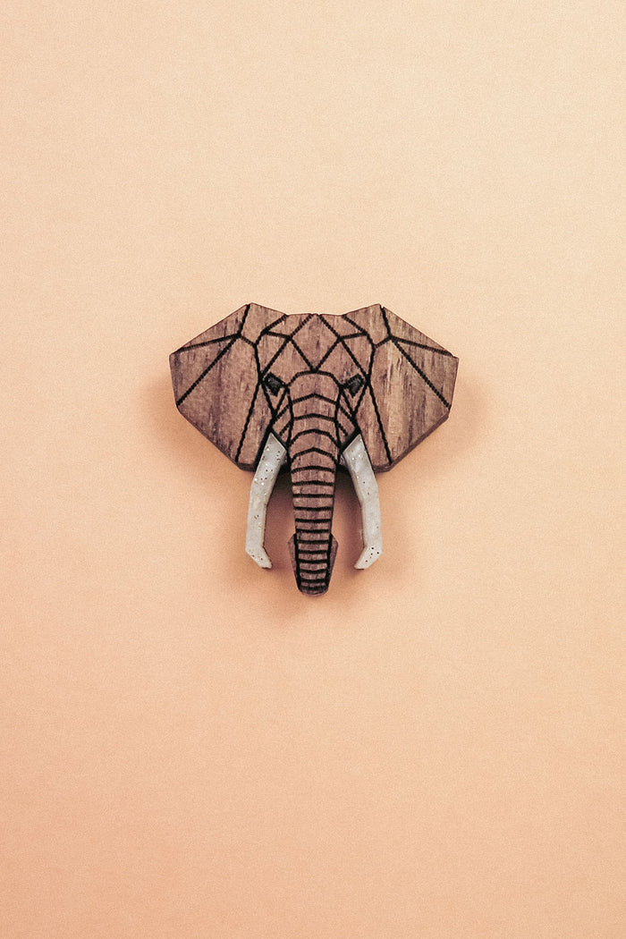 A handcrafted Elephant Pin showcases a geometric wooden carving of an elephants head with white tusks, centered on a light peach background.