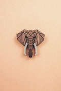 A handcrafted Elephant Pin showcases a geometric wooden carving of an elephants head with white tusks, centered on a light peach background.
