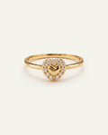 A gold heart-shaped ring featuring a central gold heart surrounded by a halo of small clear stones, set on a plain light beige background.