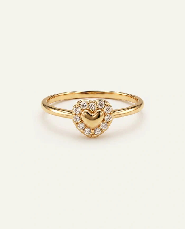 A gold heart-shaped ring featuring a central gold heart surrounded by a halo of small clear stones, set on a plain light beige background.