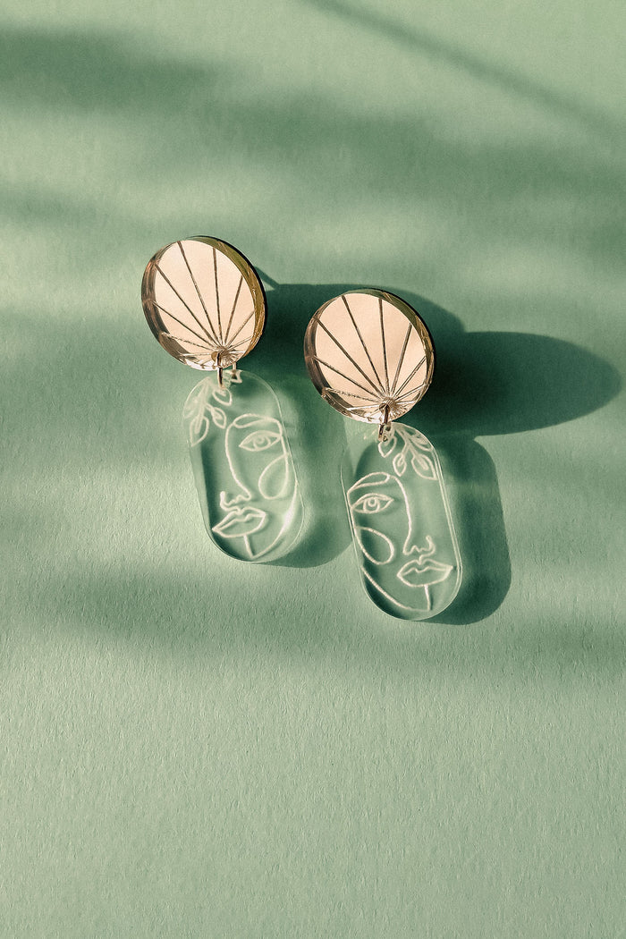 Elevate your style with these handcrafted Face Earrings: modern designs featuring round tops and dangling clear pieces etched with minimalist abstract faces, exuding artful sophistication on a green surface with soft shadows.