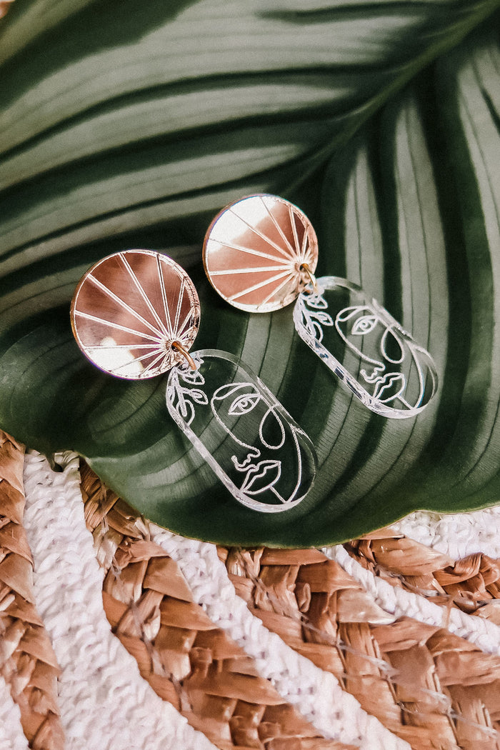 A pair of handcrafted Face Earrings with gold circular tops and abstract wire faces hang below, displayed on a green tropical leaf with a woven surface underneath to elevate your style with artful sophistication.