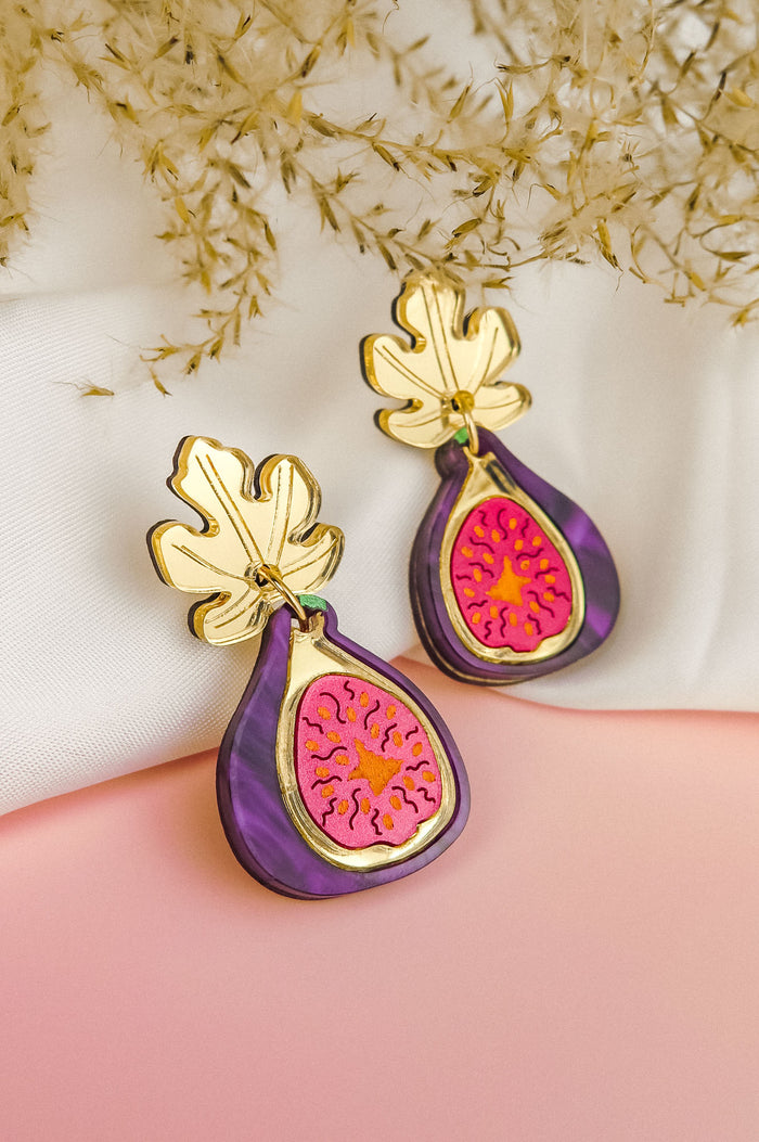 A pair of handcrafted Fig Earrings with gold mirror leaves, purple edges, and a pink center, displayed on a soft white and pink background with dried flowers.