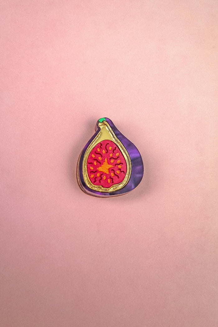 A handcrafted Fig Pin shaped like a halved fig, featuring a purple outer edge, gold outline, and vibrant red and yellow seeds, makes a lightweight accessory for any jacket or bag. Displayed on a pink background.