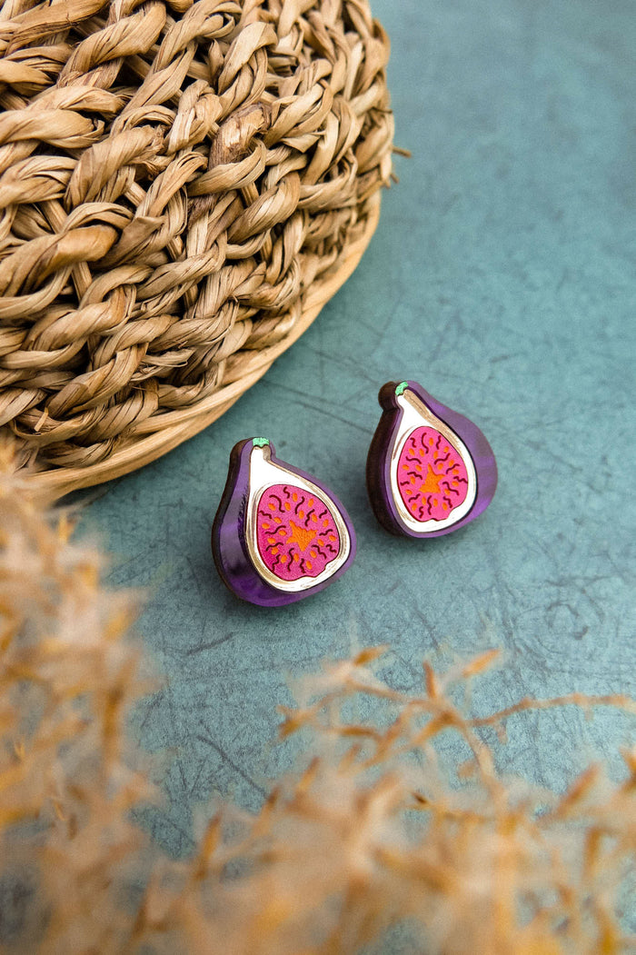 A pair of fig-shaped, hand-painted earrings with purple and pink detailing are displayed on a green surface next to a woven basket. Blurred beige plant stems are visible in the foreground.