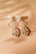 A pair of handcrafted dangling earrings with white floral studs, clear floral middle segments, and translucent, petal-shaped drops featuring cut-out and gold accents, displayed on a peach-colored surface with soft shadows.