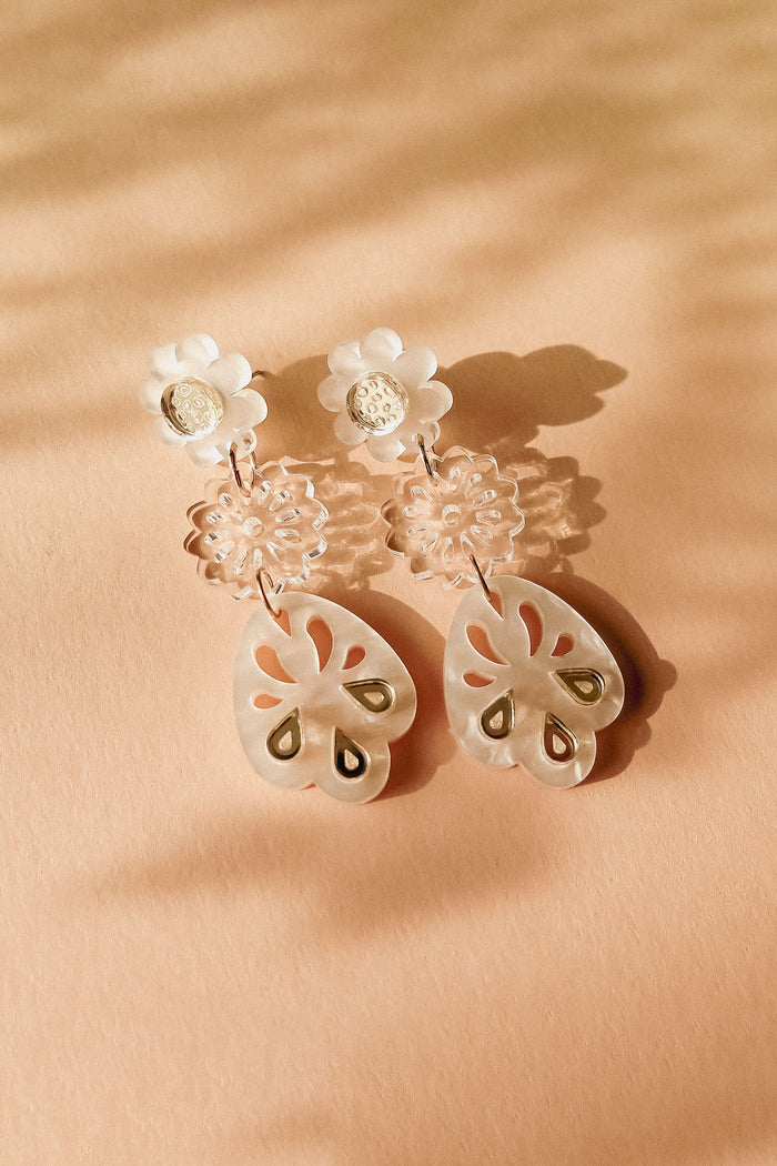 A pair of handcrafted dangling earrings with white floral studs, clear floral middle segments, and translucent, petal-shaped drops featuring cut-out and gold accents, displayed on a peach-colored surface with soft shadows.