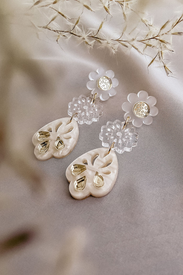 A pair of elegant, handcrafted boho earrings featuring flower-shaped clear studs and ornate, heart-shaped white pendants with gold accents, displayed on soft fabric with delicate dried flowers nearby.