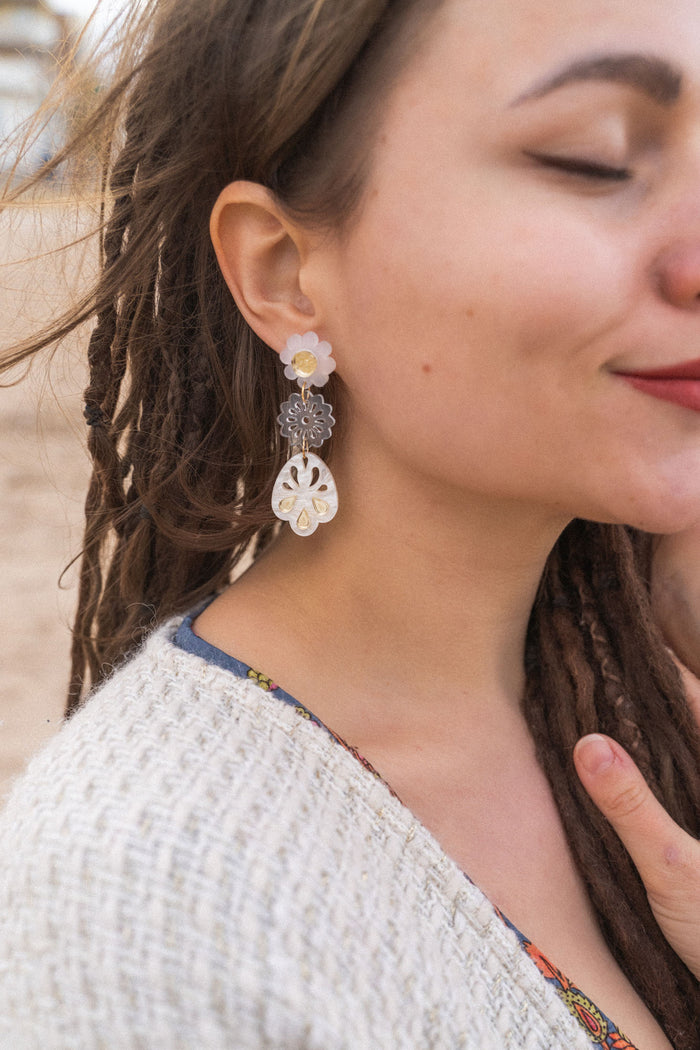 A woman with closed eyes and long brown dreadlocks wears a beige sweater and large handcrafted boho earrings, standing outdoors on a sandy beach. She smiles softly and touches her chest with one hand.