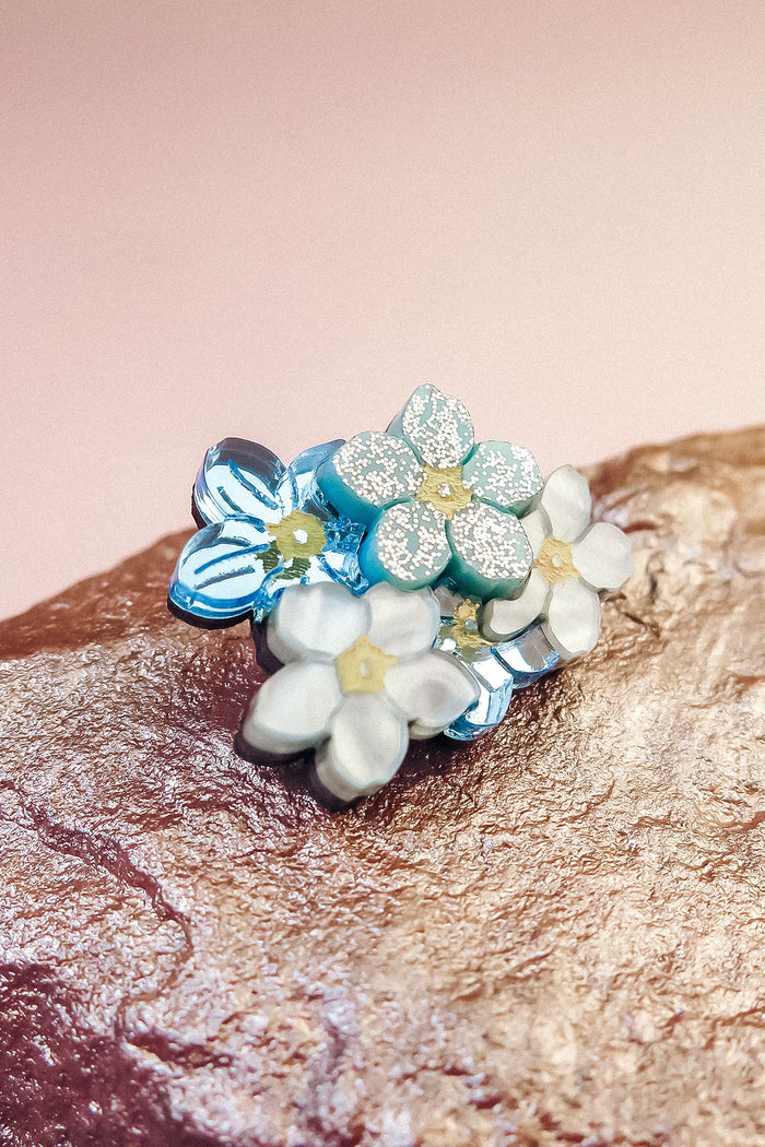 A cluster of delicate, handmade Forget Me Not Flower lapel pins with pastel blue and white petals and yellow centers rests on a textured rock against a soft pink background.