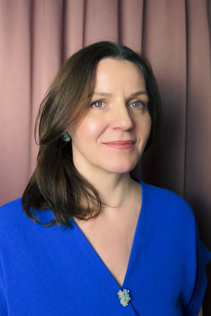 A woman with shoulder-length brown hair wearing a blue top and a handmade Forget Me Not Flower lapel pin stands in front of mauve curtains, smiling gently at the camera.