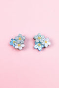 A pair of floral earrings, the Forget Me Not Studs, featuring blue, white, and glittery silver flowers are displayed on a light pink background—wearable art that brightens any look.