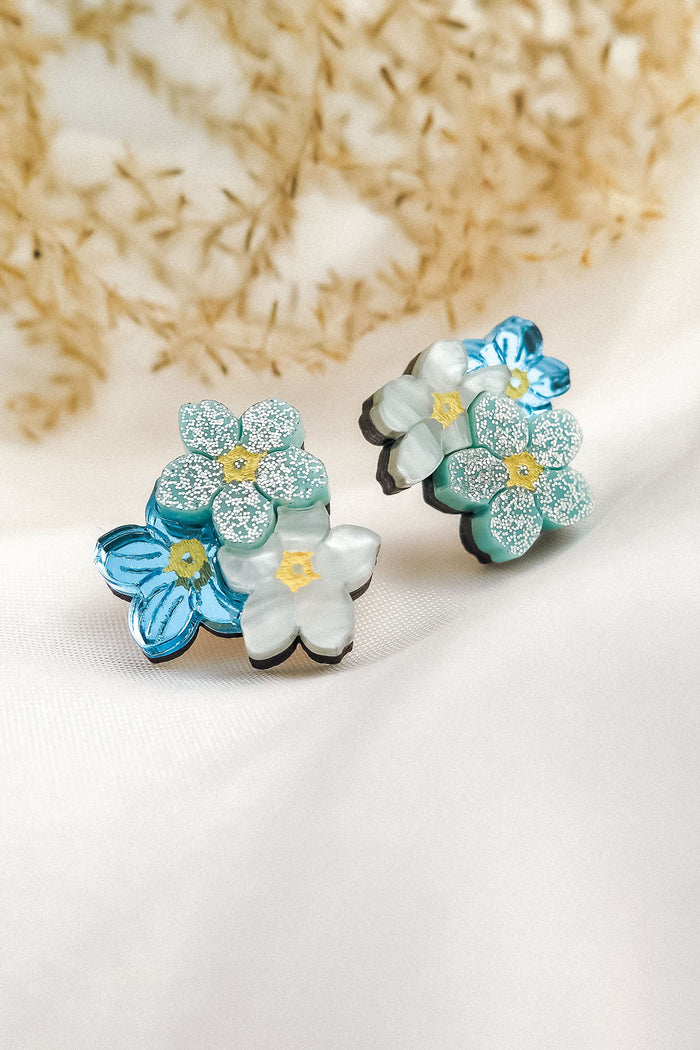 A pair of decorative Forget Me Not Studs featuring layered blue and white flowers with glittery petals, displayed on a soft white fabric with dried plants in the background—delicate floral earrings that are true wearable art.