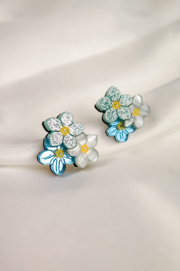 A pair of Forget Me Not Studs, featuring blue and white petals, yellow centers, and glittery accents, these floral earrings are true wearable art displayed on a soft, light-colored fabric background.