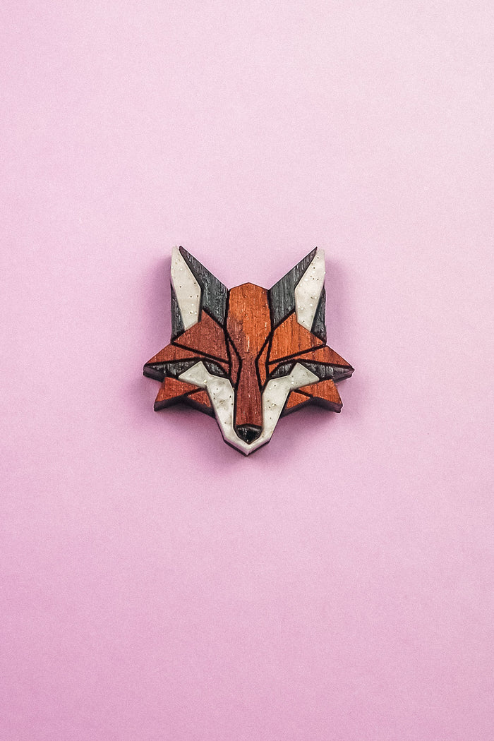 A geometric, stylized fox head brooch—handcrafted from recycled wood in shades of brown, black, and white—rests on a light pink background, making it an eco-conscious accessory with modern flair.