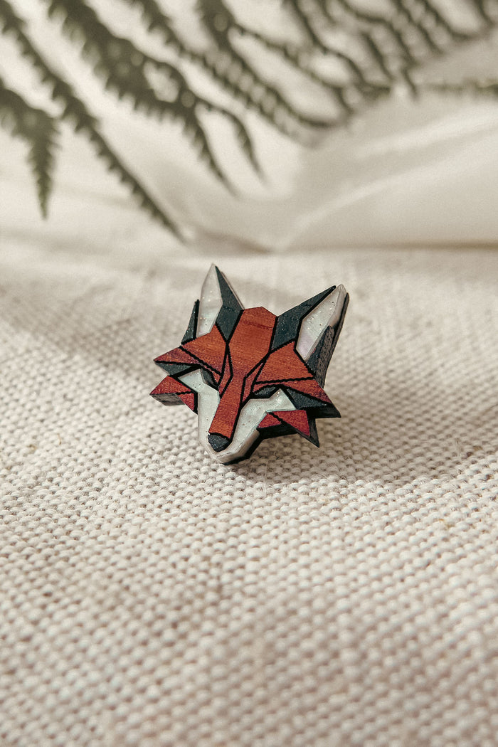 A geometric, handcrafted fox pin made from recycled wood rests on textured beige fabric, with blurred green fern leaves in the background.
