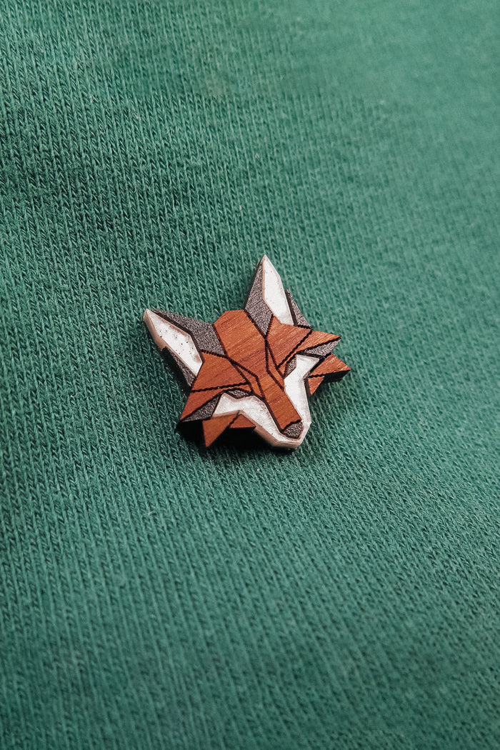 A small, geometric handcrafted fox pin made from recycled wood features orange, brown, and white sections and is pinned to a green textured fabric.