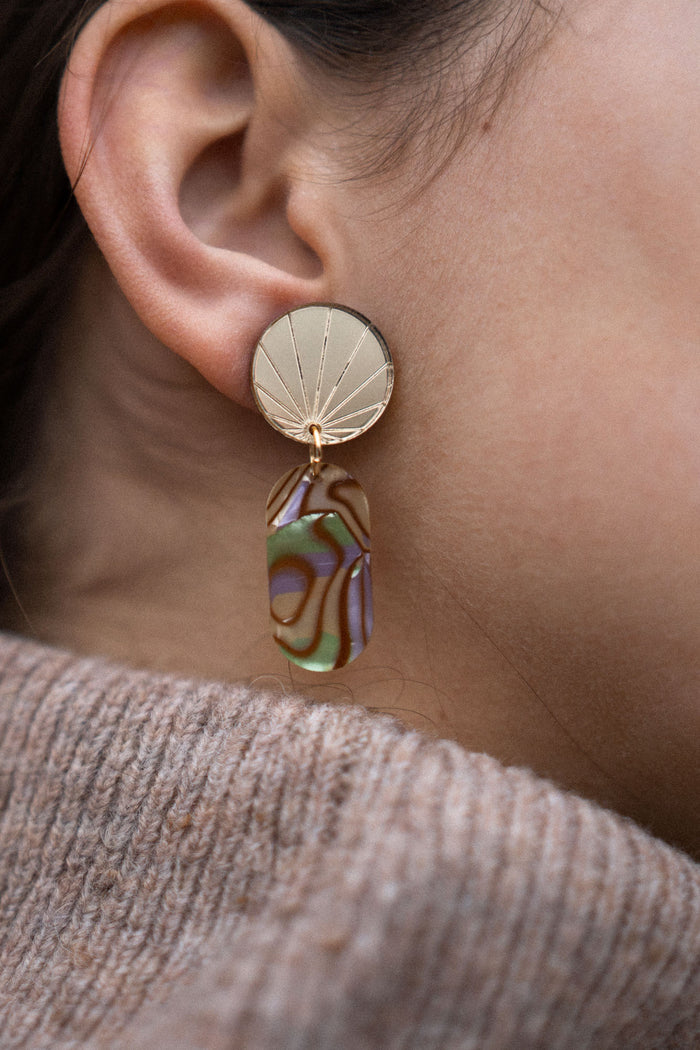 Close-up of a person’s ear wearing a handcrafted gold earring with a sunburst design, featuring a dangling abstract, multicolored abalone piece. The person is wearing a taupe knit sweater.