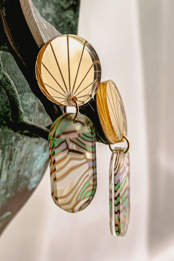 Close-up of handcrafted gold earrings with engraved fan-patterned tops and elongated oval dangles made of translucent abalone, showcasing wavy green, brown, and cream stripes.