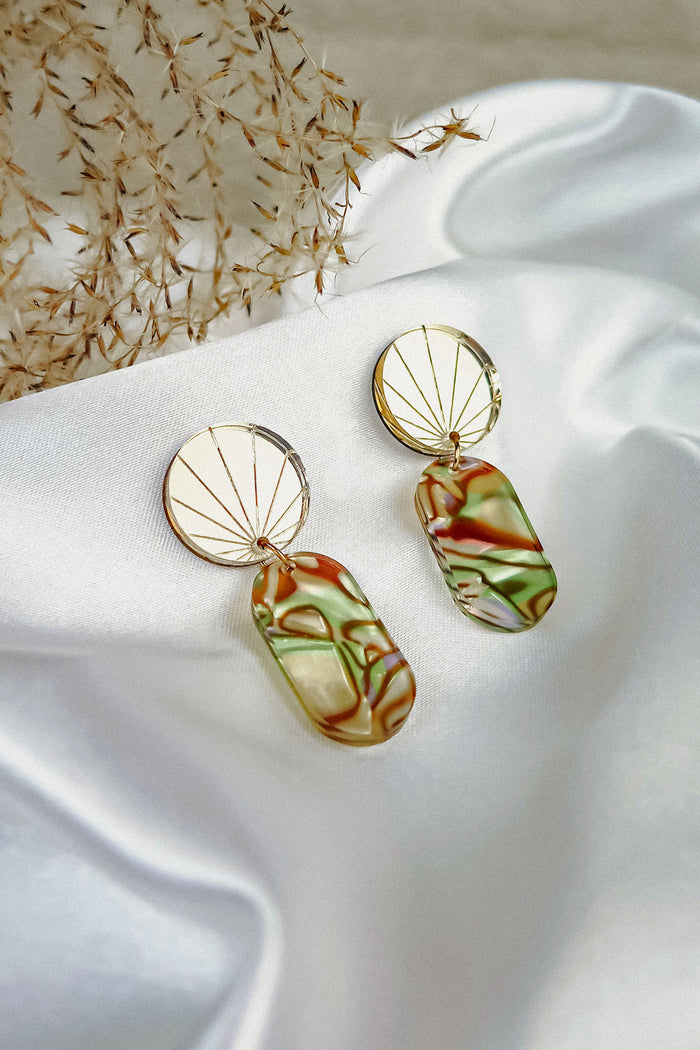Two handcrafted gold earrings rest on white satin fabric. Each features a sunburst design and a dangling rectangular pendant with marbled green, brown, and cream hues, reminiscent of abalone. Delicate dried foliage lies nearby.