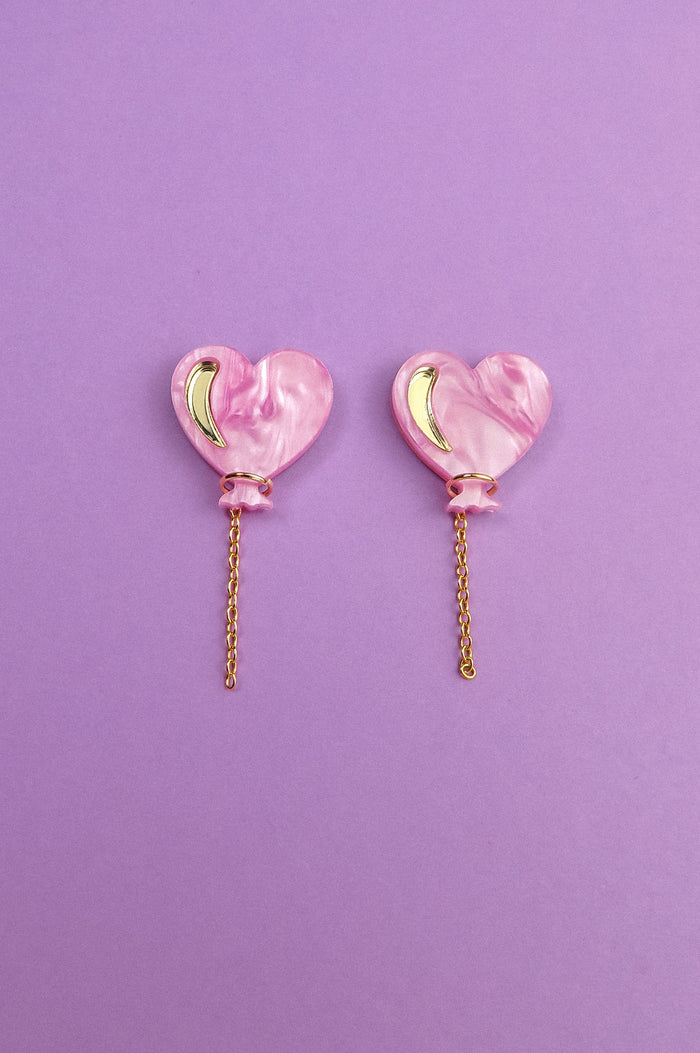A pair of Heart Balloon Studs—pink, heart-shaped earrings with gold crescent accents and dangling gold chains—adds vibrant style, displayed on a purple background.