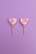 A pair of Heart Balloon Studs—pink, heart-shaped earrings with gold crescent accents and dangling gold chains—adds vibrant style, displayed on a purple background.