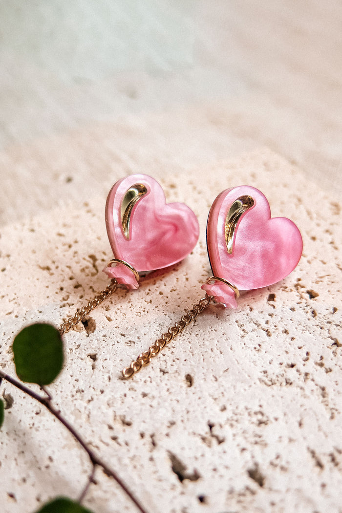 A pair of pink Heart Balloon Studs with gold accents and dangling gold chains, displayed on a textured, light-colored stone surface. A small green plant is visible in the lower left corner, completing this vibrant style.