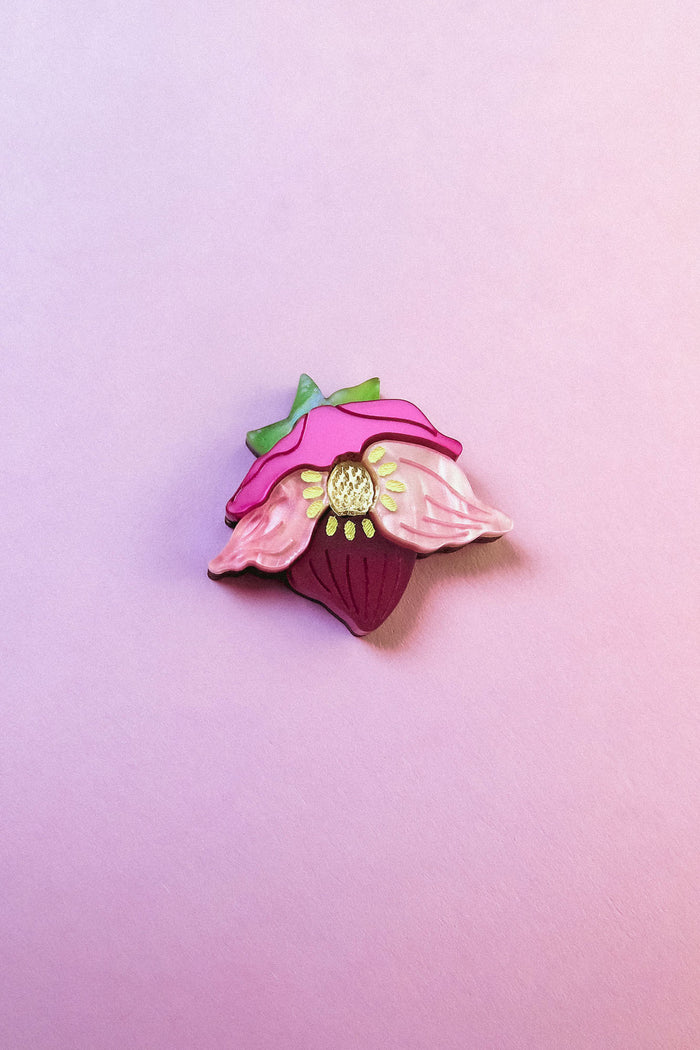 A colorful enamel lapel pin shaped like a strawberry with pink petals and green leaves is displayed on a soft pink background—perfect for handmade accessory lovers or fans of the unique purple hellebore flower.