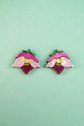 Two colorful Hellebore Stud Earrings, shaped like stylized pink flowers with green leaves and yellow centers, are displayed on a light turquoise background—perfect handcrafted floral jewelry for unique accessories lovers.