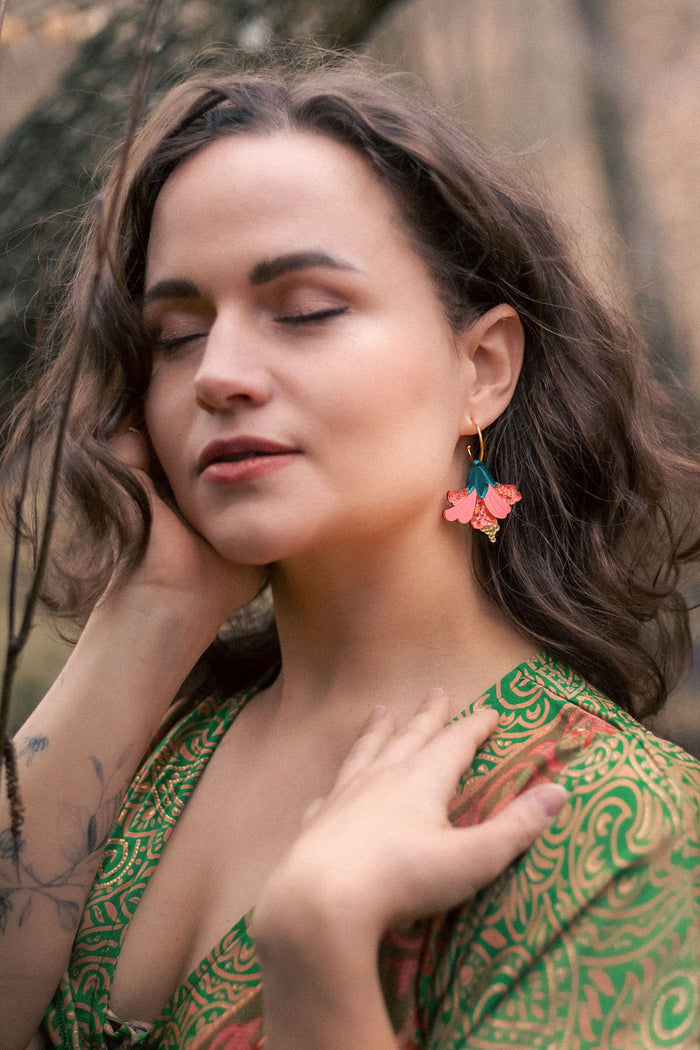 A woman with wavy brown hair and closed eyes stands outdoors, wearing a green patterned dress and vibrant Hibiscus Earrings, touching her face and neck gently, with a tattoo visible on her forearm.