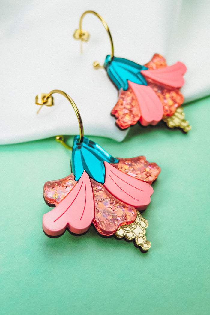 Vibrant Hibiscus Earrings with gold hooks, featuring colorful pink, blue, and glittery accents, make a bold tropical statement against a striking green and white background.