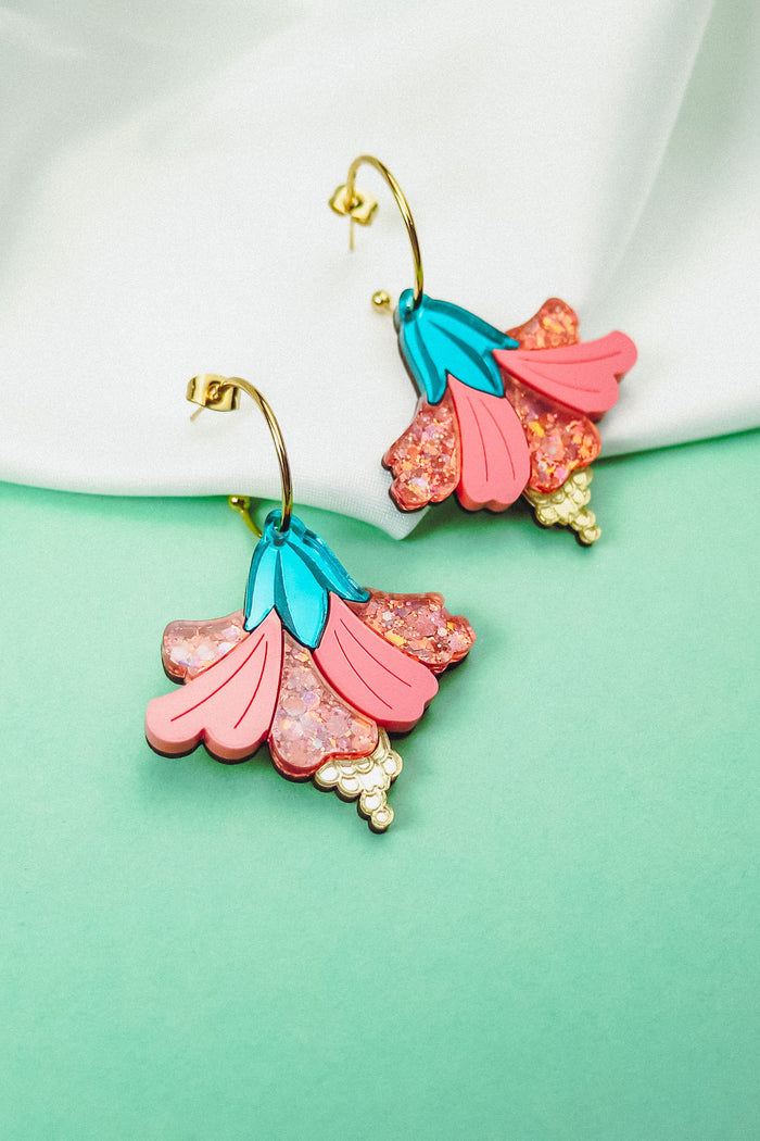 A vibrant pair of Hibiscus Earrings with gold hooks, featuring pink and blue petals and sparkling pink accents, make a tropical statement against a white cloth and mint green background.