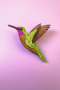A handcrafted Hummingbird Brooch with vibrant colors—green, pink, brown, and gold glitter accents—shines beautifully against a light pink background.