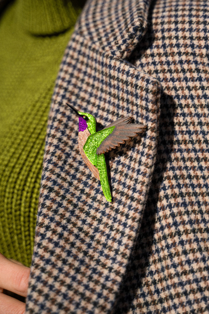 A close-up of a handcrafted hummingbird brooch, made from premium materials, pinned to a houndstooth-patterned coat with a green turtleneck sweater visible underneath.