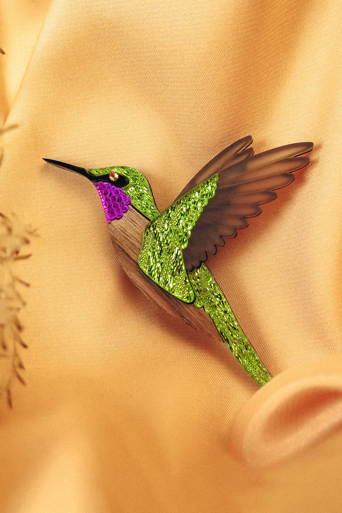 A handcrafted hummingbird brooch with vibrant green and purple glittery hues and brown wings rests on soft, peach-colored fabric.