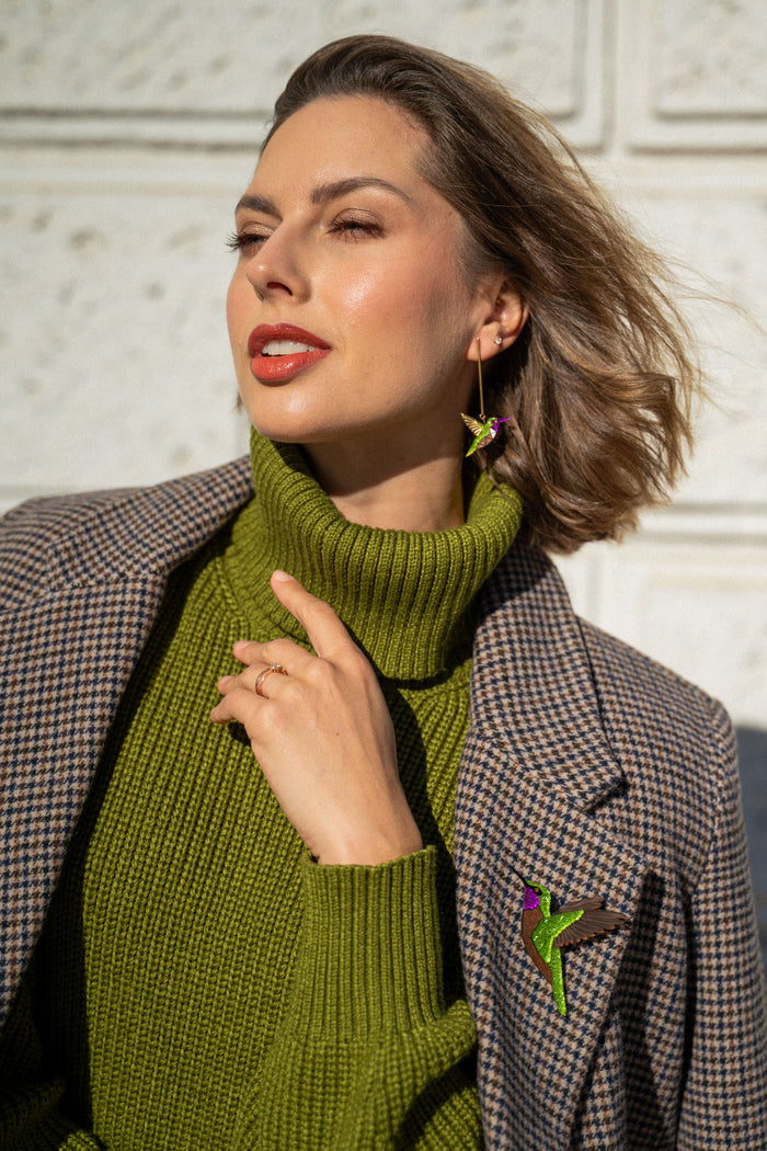 Woman wearing a green turtleneck sweater and plaid coat, accessorized with handcrafted Hummingbird Earrings and a brooch, poses outdoors against a light textured wall, touching her collar and looking off to the side.