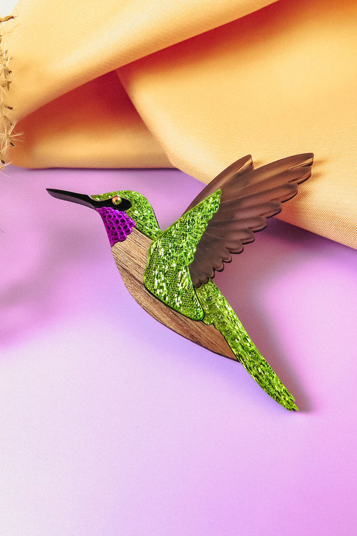 A handcrafted Hummingbird Brooch with vibrant colors—glittery green wings, a purple head, and brown accents—rests on a pastel pink surface with a yellow fabric backdrop.
