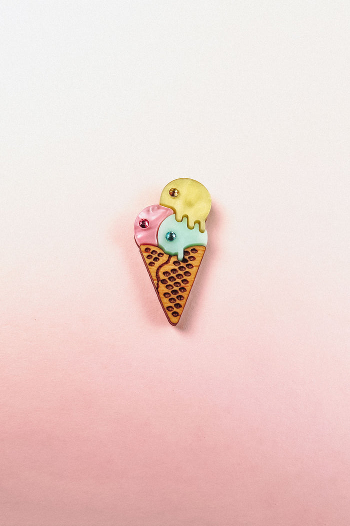 A playful design, this handcrafted Ice Cream Pin features a colorful enamel cone with three scoops—pink, blue, and yellow—set against a soft pink and white gradient background.