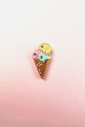 A playful design, this handcrafted Ice Cream Pin features a colorful enamel cone with three scoops—pink, blue, and yellow—set against a soft pink and white gradient background.