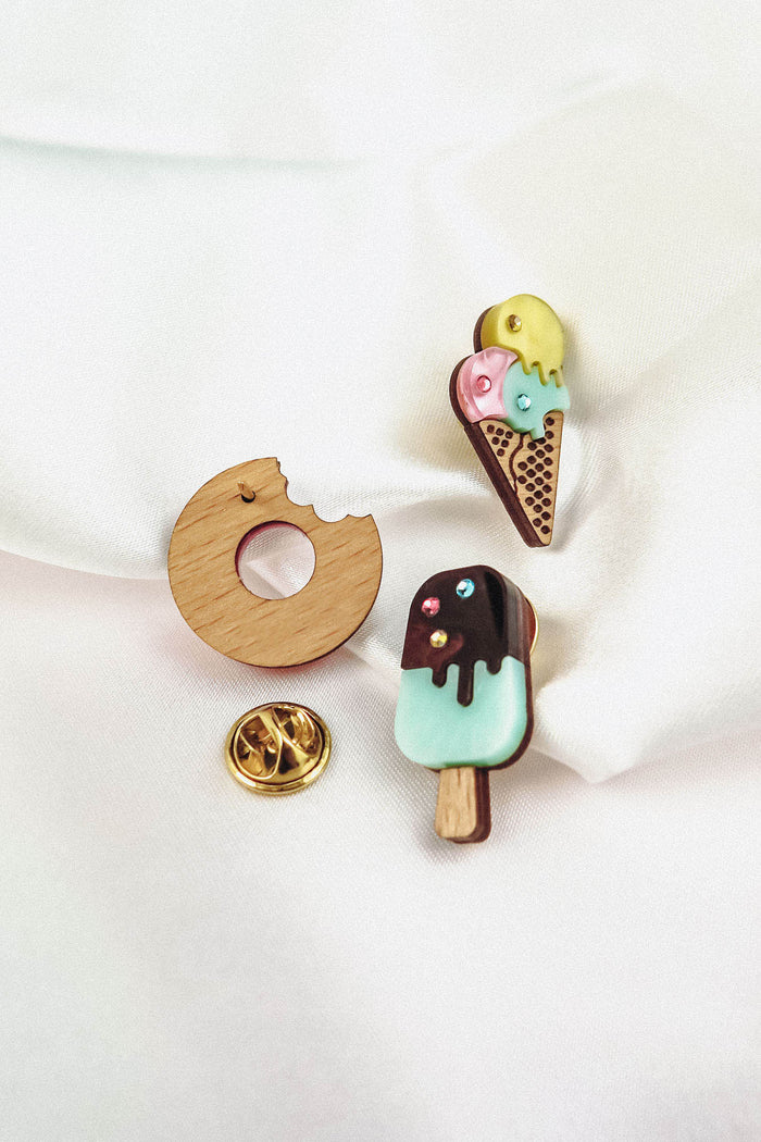 Three handcrafted enamel pins on white fabric: a Donut Pin with a bite taken out, a triple scoop ice cream cone, and a chocolate-covered popsicle, each sparkling with rhinestones and paired with a gold pin backing nearby.